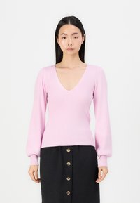 Suéter rosa com nervuras, decote em V profundo, mangas bufantes longas e cintura justa, combinado com uma saia preta de botões.