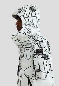 Profil osoby w białej kurtce z kapturem ozdobionej czarną abstrakcyjną grafiką linii, wzorami tekstowymi i naszywką na rękawie.