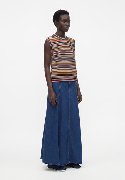 Femme portant un haut sans manches rayé multicolore et une longue jupe en jean bleue, debout devant un fond blanc uni.
