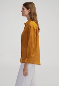 Senfgelbe, halb-transparente Button-Down-Bluse mit langen Ärmeln, strukturiertem Stoff und lockerer Passform, kombiniert mit weißen Hosen.