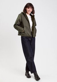 Veste bomber vert olive avec une doublure en fausse fourrure, fermeture éclair à l'avant et poches latérales, associée à un jean droit en denim foncé et des bottines noires.