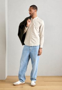 Man in een beige overhemd met knopen en lichtblauwe spijkerbroek houdt een donkere jas over zijn schouder terwijl hij op een houten vloer staat tegen een grijze muur.