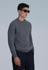 Pull en tricot gris avec texture côtelée et rayures verticales, présentant un col rond et des manches longues, porté avec un pantalon sombre et des lunettes de soleil.