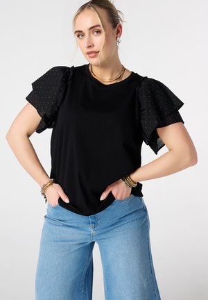 Vrouw draagt een zwarte top met doorschijnende, geplooide polkadotmouwen en lichtblauwe spijkerbroek, staat met handen in de zakken tegen een effen achtergrond.