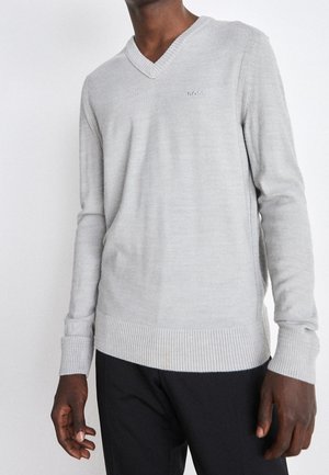 Homme portant un pull col V gris clair avec le logo BOSS sur la poitrine et un pantalon noir sur fond blanc.