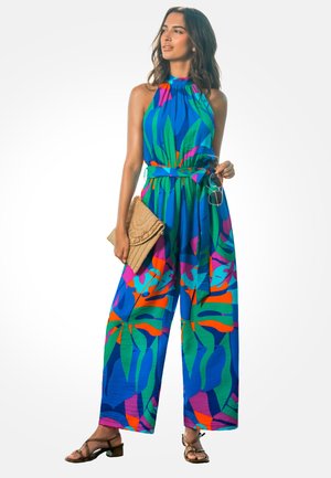 Kobieta ubrana w jaskrawy, wielokolorowy kombinezon na ramiączkach, trzymająca plecioną kopertówkę i okulary przeciwsłoneczne, pozująca na jednolitym białym tle.