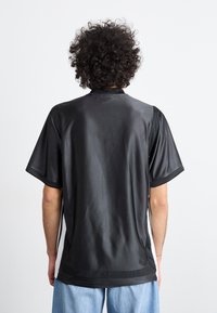 Camisa preta de mangas curtas com uma textura suave e brilhante. Apresenta um colarinho simples e painéis de malha ao longo das laterais.