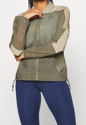 Person trägt eine grün-beige Jacke mit Reißverschluss und blaue Leggings, Arme verschränkt, eine Hand berührt das Kinn.