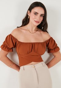 Rustrød off-shoulder top med pufærmer, tekstureret stof og tætsiddende design; parret med lyse beige høj talje bukser.