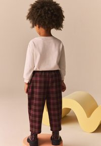 El niño lleva una camiseta de manga larga gris claro y unos pantalones cuadros burdeos con un corte relajado. Unos zapatos negros con cordones completan el conjunto.