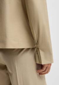 Beige sporttakk med rund krage. Har en knappdetalj ved mansjetten og en subtil tekstur, vist sammen med matchende bukser.