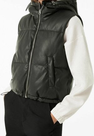 Person trägt eine schwarze gesteppte ärmellose Lederweste mit Frontreißverschluss über einem weißen Langarmhemd, Hände in den Taschen einer schwarzen Hose.