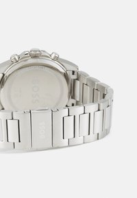 Montre en acier inoxydable argenté avec un fond de boîtier lisse, trois couronnes, fonction étanche et un bracelet à maillons affichant le nom de la marque "BOSS."