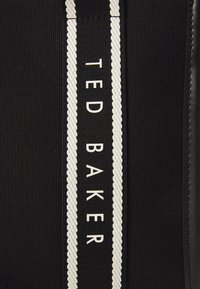 Zwarte stoffen tas met een opvallende witte gevlochten streep en de merknaam "TED BAKER" in hoofdletters. Glad textuur.