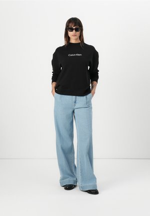 Felpa nera con logo "Calvin Klein", abbinata a jeans a gamba larga blu chiaro. Il modello indossa occhiali da sole neri e si trova in piedi con le mani in tasca.