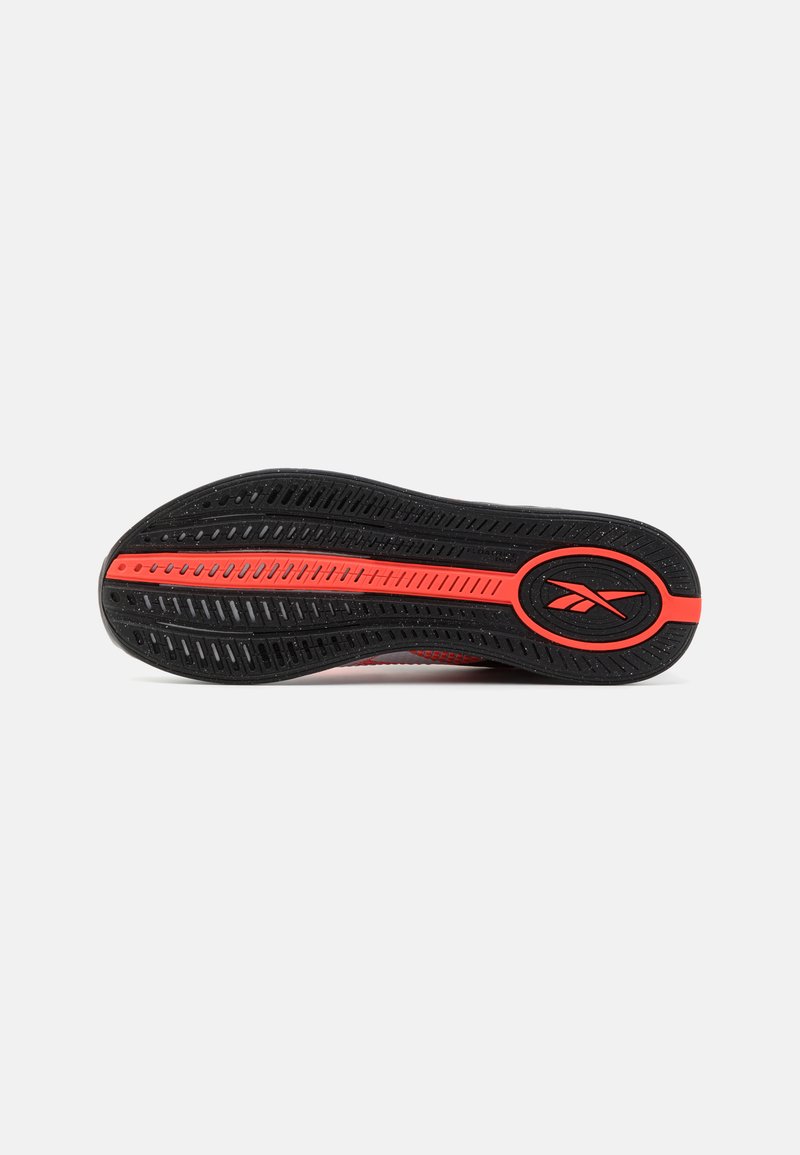 Suela de zapato de goma negra con una línea roja y un logo ovalado. Presenta patrones de agarre texturizados y orificios de ventilación para tracción.