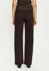 Jeans larghi marroni mostrati da dietro, con due tasche a toppa, cuciture a contrasto e una consistenza di cotone pesante e liscia.