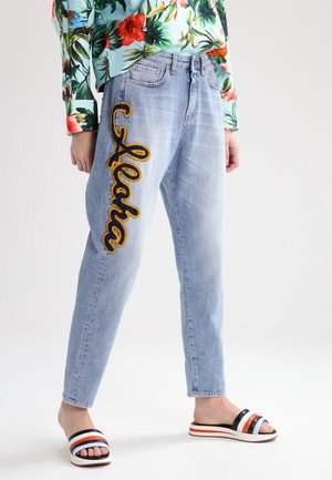 Vaqueros azul claro con un gran bordado amarillo y negro de "Alaska" en la pierna izquierda, combinados con una blusa floral y sandalias tipo slip-on a rayas.