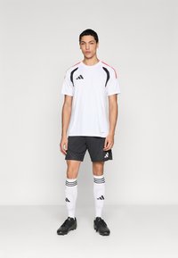 Λευκό αθλητικό πουκάμισο με μαύρες και κόκκινες λεπτομέρειες, συνδυασμένο με σκούρα γκρι σορτς, μαύρα ποδοσφαιρικά παπούτσια και λευκές ποδοσφαιρικές κάλτσες μέχρι το γόνατο με μαύρες ρίγες.