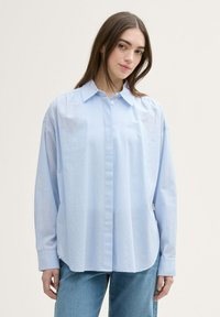 Camisa de botones azul claro con un corte relajado, de mangas largas y cuello puntiagudo. Fabricada con una tela suave y de textura sutil.