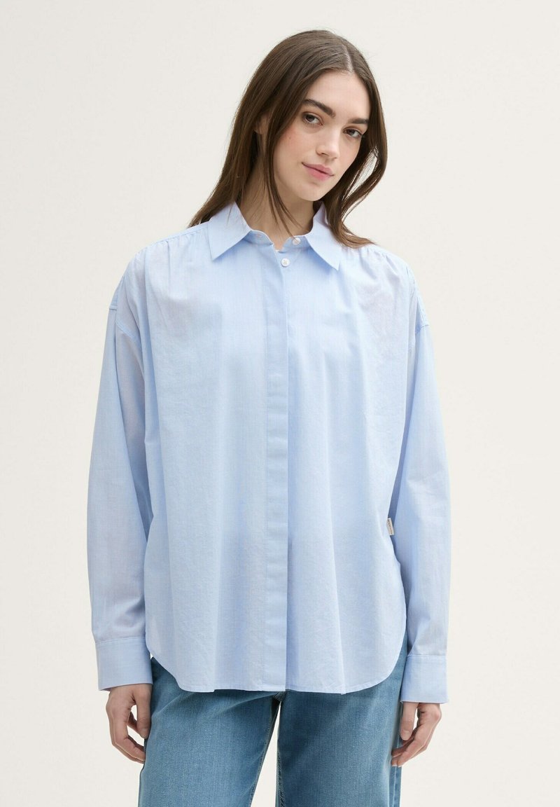 Camisa de botones azul claro con un corte relajado, de mangas largas y cuello puntiagudo. Fabricada con una tela suave y de textura sutil.