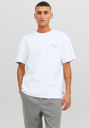 Camiseta blanca de manga corta con cuello redondo y texto impreso en el lado izquierdo del pecho. Material de algodón suave, corte relajado. Combinada con pantalones grises.