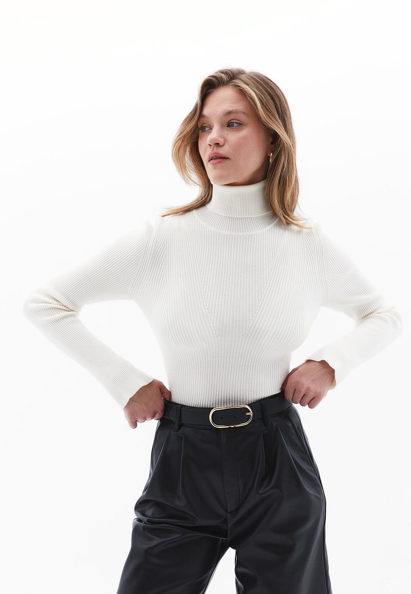 Weißer gerippter Rollkragenpullover mit langen Ärmeln, der eine figurbetonte Silhouette bietet. Kombiniert mit hochtaillierten schwarzen Hosen und einem goldenen Gürtel.