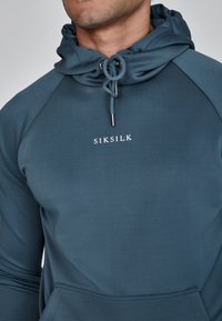 Dark teal hoodie with a drawstring hood, featuring a kangaroo pocket and white "SIKSILK" logo on the chest. Smooth fabric texture.