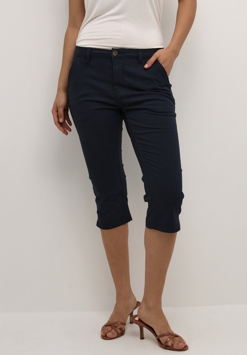 Femme portant un pantalon court bleu marine avec bouton sur le devant et poches, associé à des sandales à talons marron et un top blanc sans manches.