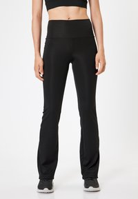 Pantalones deportivos negros con una cintura alta, silueta ajustada, tejido ligero y piernas ligeramente acampanadas, combinados con zapatillas grises.