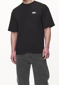 Camiseta de algodón negra con cuello redondo y mangas cortas, que presenta un logo blanco en el pecho izquierdo. Combinada con jeans grises.