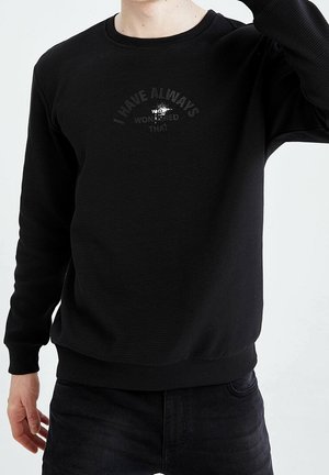 Mand iført en sort ribbet sweatshirt med svag tekst "I have always wanted that" i mørkt tryk på brystet, kombineret med sorte jeans.