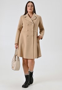 Cappotto beige doppiopetto con colletto e tasche frontali. Tessuto texturizzato, bottoni in tonalità argento, abbinato a stivaletti neri con lacci e una borsa beige.