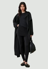Conjunto negro en capas que incluye un abrigo largo, una blusa ajustada, leggings y zapatos sin cordones con acentos dorados. Textura suave y diseño monocromático.