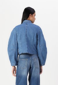 Giacca in denim blu tagliata, con maniche lunghe e larghe e un design della cucitura posteriore strutturato, abbinata a jeans blu a vita alta.