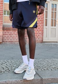 Sötétkék sportnadrág sárga és rózsaszín részletekkel, fehér bokazoknival párosítva, valamint bézs sportcipő, kék logóval és textúrázott dizájnnal.