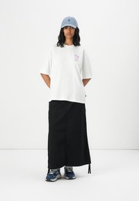 Vit oversized t-shirt med rosa grafik på bröstet, kombinerat med en lång svart kjol och blå sneakers. En ljusblå keps fulländar outfiten.