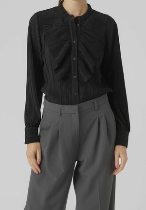 Czarna plisowana bluzka z falbanami z przodu, zapinana na guziki, z wysokim kołnierzem. Noszona z dopasowanymi szarymi spodniami. Gładka faktura tkaniny.