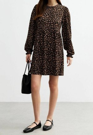 Woman wearing a long-sleeve, knee-length leopard print dress, holding a black handbag, and wearing black Mary Jane flats.