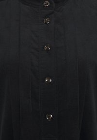 Camicia in corduroy nero caratterizzata da un colletto alto, pattina frontale con sei bottoni scuri e linee di texture verticali. Tessuto morbido con una finitura liscia.