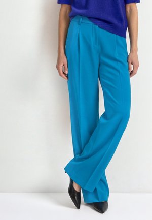 Femme portant un pantalon large bleu vif, un pull bleu foncé et des chaussures noires à bout pointu, debout les jambes croisées.