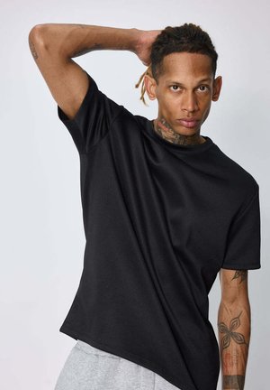 Homme tatoué portant un t-shirt noir et un pantalon gris, pose une main derrière la tête devant un fond uni clair.