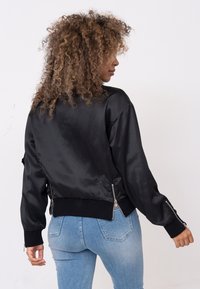 Zwarte satijnen bomberjack met ribgebreide boorden en zoom. Heeft ritszakken aan de zijkanten en een ontspannen pasvorm. Glad tekstuur.