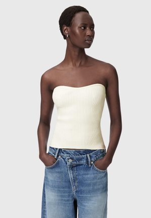 Vrouw met kort haar, een strapless geribd wit topje en losse blauwe jeans met handen in de zakken, kijkt zijwaarts.