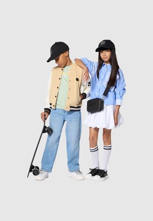 Deux enfants en vêtements de rue décontractés portant des casquettes noires DKNY ; l'un tient un skateboard, l'autre porte un sac en bandoulière et des chaussettes montantes.