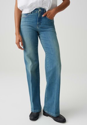 Persona che indossa jeans azzurri a gamba dritta e mocassini neri, con una mano in tasca e camicia bianca infilata.