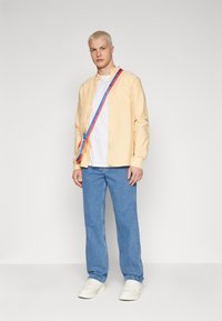 Light yellow button-up shirt over a white t-shirt, paired with blue jeans and white sneakers. A colorful striped bag crosses the body.