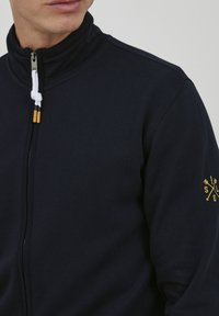 Felpa navy con zip e colletto alto, realizzata in un morbido tessuto. Presenta un cordino bianco con punte in legno e un logo giallo sulla manica.