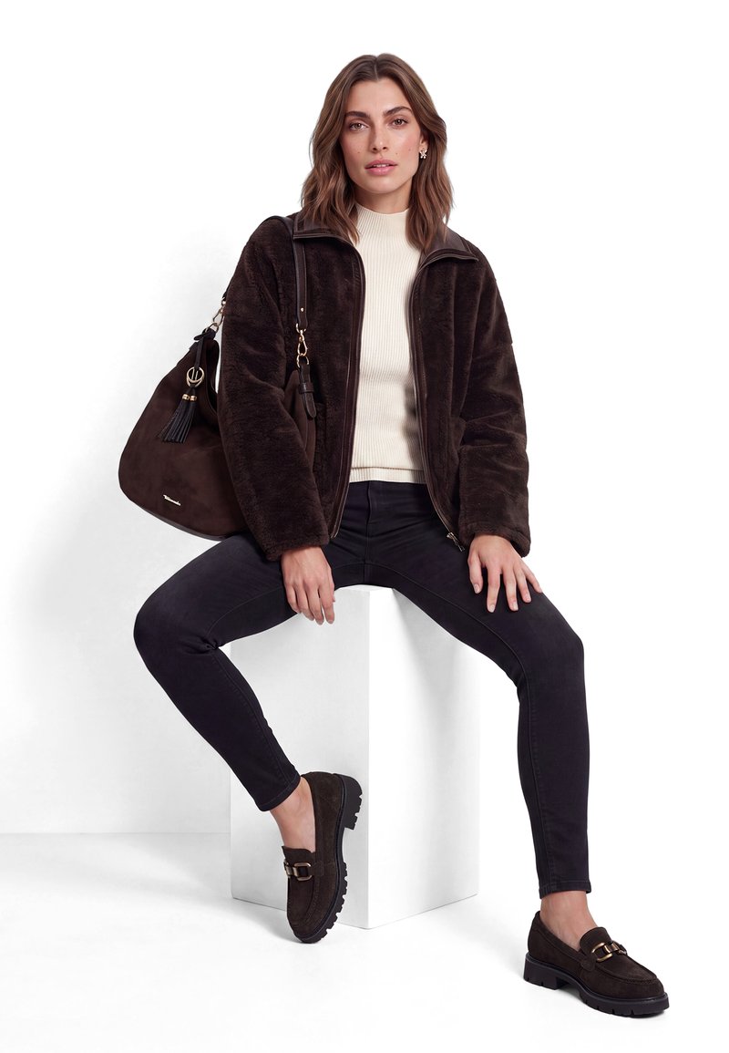 Femme portant une veste duveteuse marron foncé, un pantalon noir, des mocassins foncés et un pull col roulé crème, assise sur un cube blanc avec un sac marron.