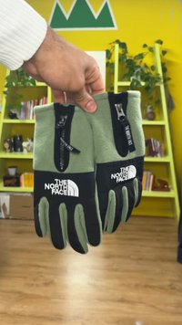 Zelene flis rokavice s črnimi poudarki in zadrgami, z logotipom "The North Face" jasno prikazanim na notranji strani dlani in na hrbtni strani.